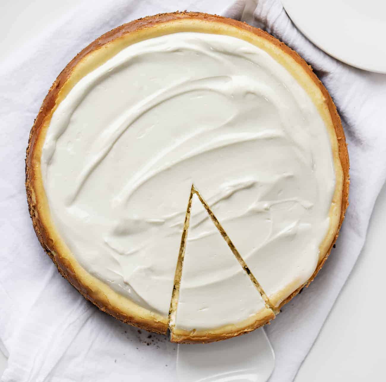 Overhead Image of Brownie Bottom Cheesecake Recipe with Sour Cream Frosting Topping with One Piece Cut and Being Removed with Pie Server