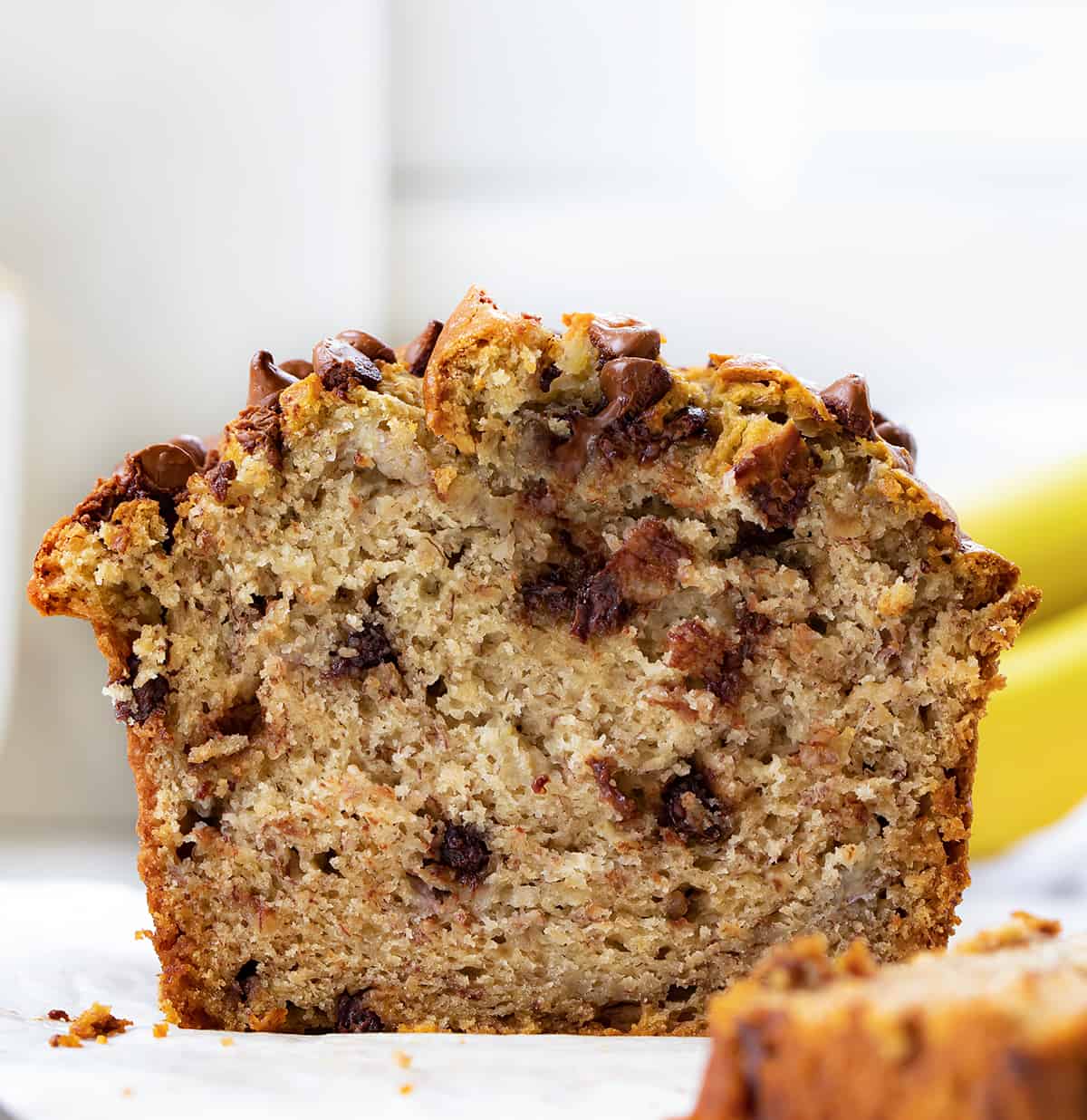 Cut into Loaf of Chocolate Chip Banana Bread on a White Counter with Bananas in the Background.