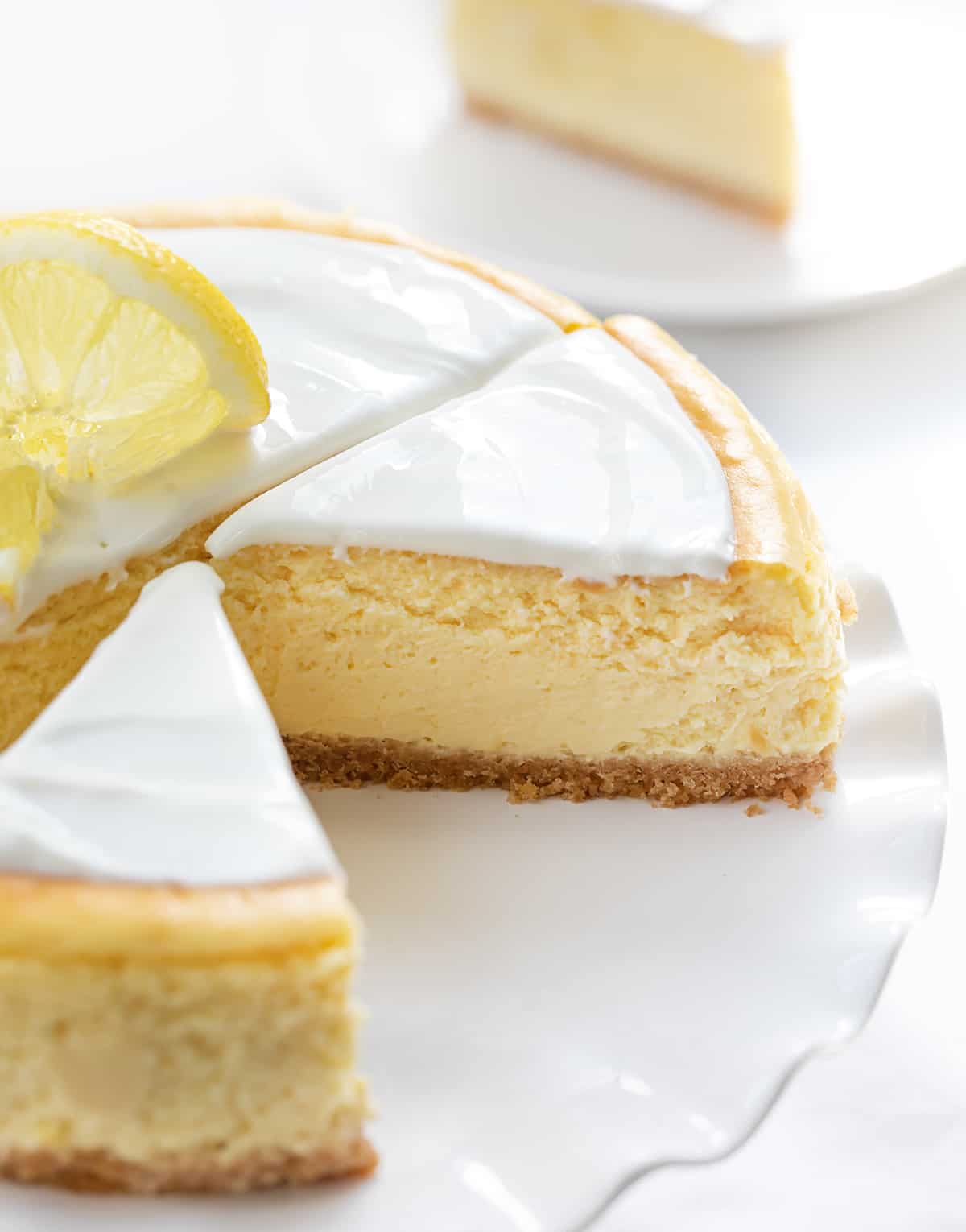 One Lemon Cheesecake on a White Cake Stand with Ruffles and Pieces Cut with a Piece on a Plate in the Background.