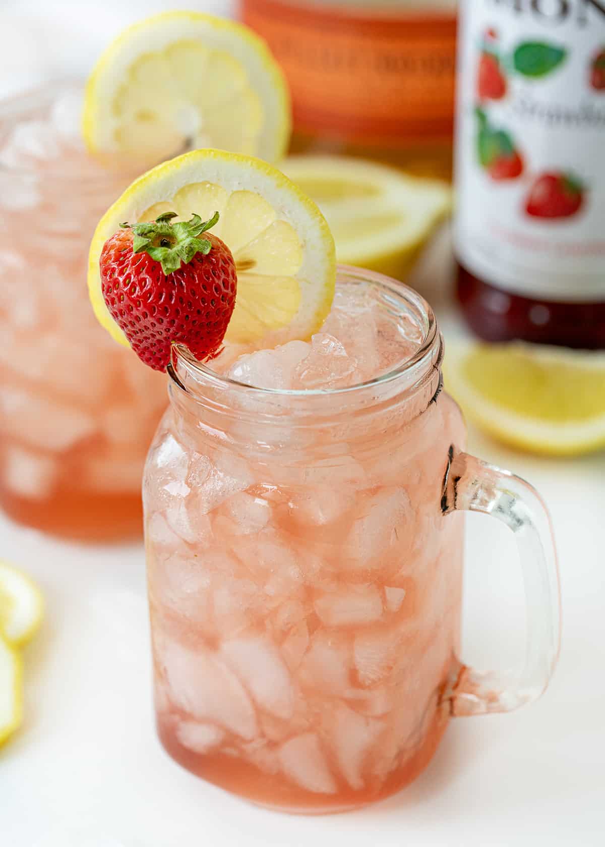 Glasses of Whiskey Strawberry Lemonade on a White Counter with Strawberry Syrup in the Background.